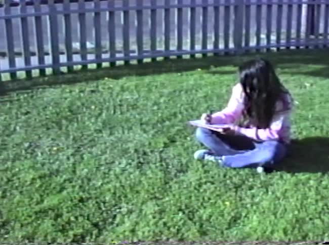 A young person with long dark hair sits crosslegged on a sunny patch of grass focused on writing or drawing on a clipboard they are positioned in a yard enclosed by a white picket fence with a street and parked cars visible in the background