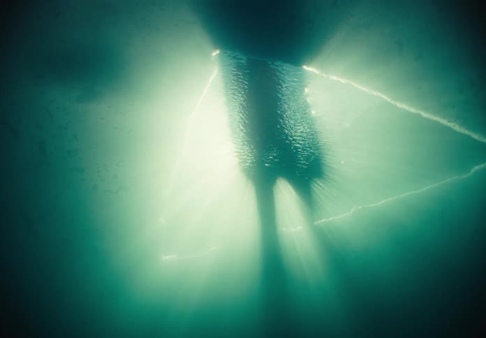A view from under the water shows the dark shape of a person standing on a surface as bright sunlight beams through a triangular opening and creates glowing rays in the green murky depths