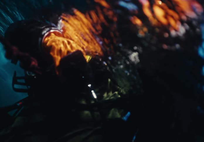 A diver wearing orange gloves and a dark suit pulls a red rope while submerged in deep blue water near a bright ice shelf