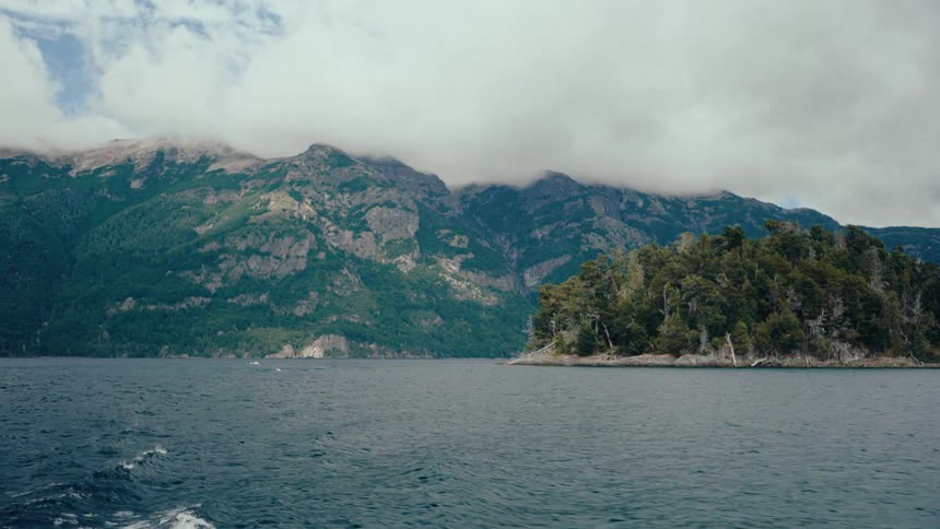 A view from a boat shows dark blue water and a forested island with large mountains and white clouds in the background