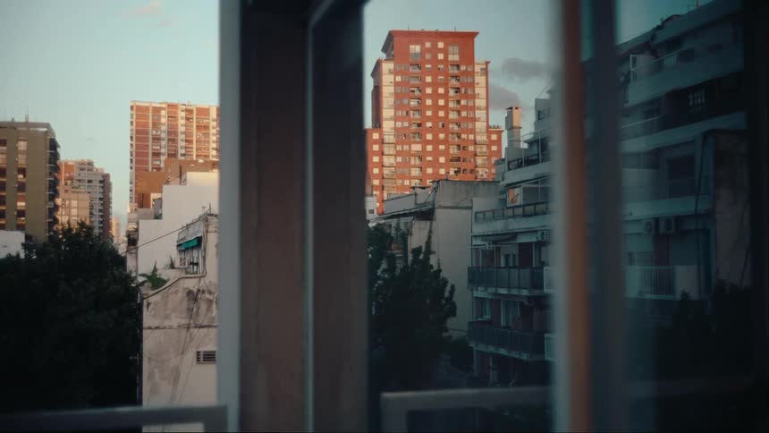 A view of sunlit apartment buildings in a cityscape framed by the vertical edges of a nearby window and balcony railing
