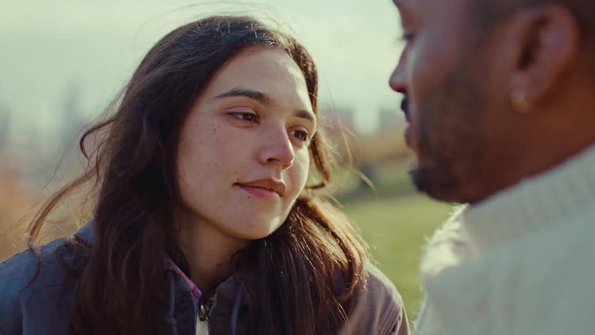 A woman with long brown hair looks at a man with a beard while they stand outside in a park on a sunny day during a close view