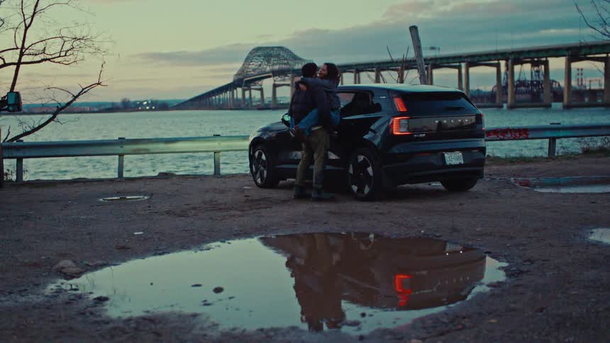 Couple hugging beside a parked car near a bridge over the river at dusk