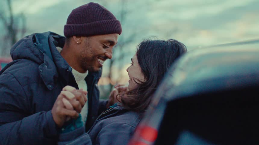Young adult man holding and playfully kissing a young adult woman against a car with trees on the background outdoors during dusk or dawn