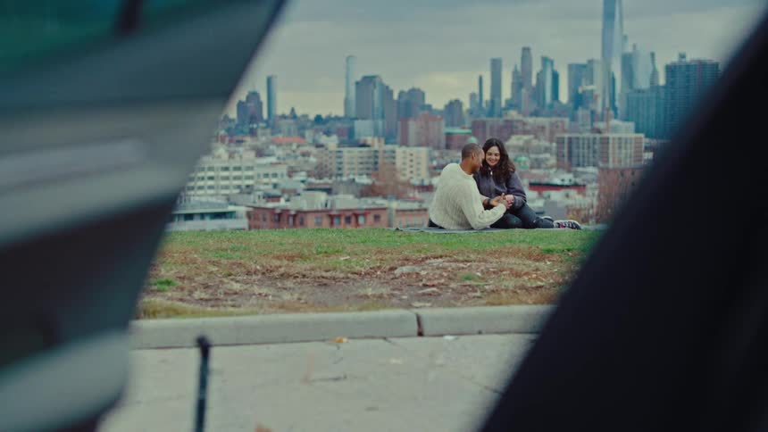 Couple sits on a blanket on a hill overlooking the city while their car sits nearby with an open car door