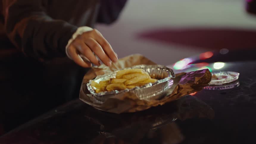 A person reaches for a french fry from a foil container sitting on top of a dark car at night with colorful lights reflecting on the surface