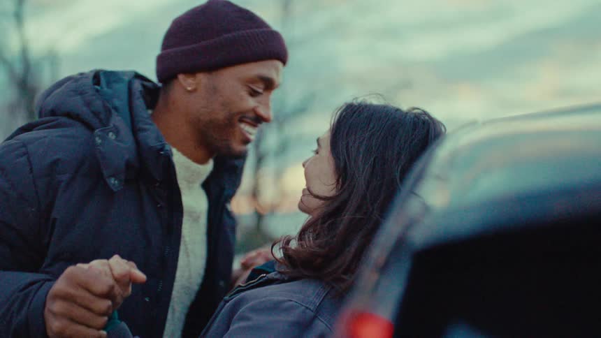 A young couple stand outdoors near a vehicle holding hands and kissing
