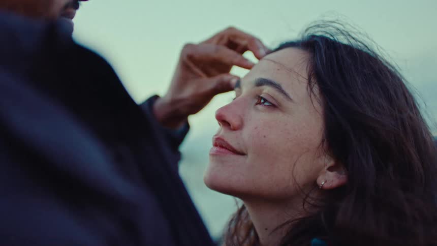 A man gently brushes hair away from the forehead of a smiling woman while they look at each other during a close view outdoors at dusk