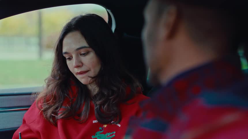 A young woman in a red sweatshirt sits in a car gazing thoughtfully at a man in the foreground during a quiet conversation