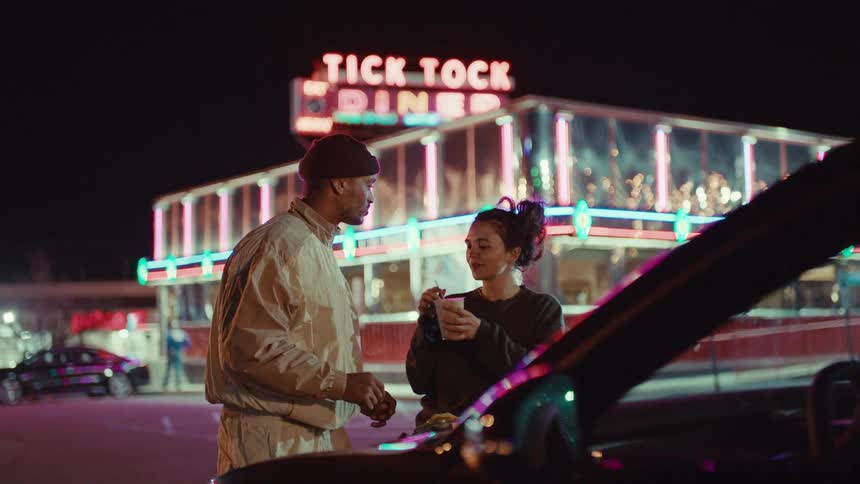 Laughing young adult couple talking and eating on the parking lot of a diner next to a car outdoors during night time