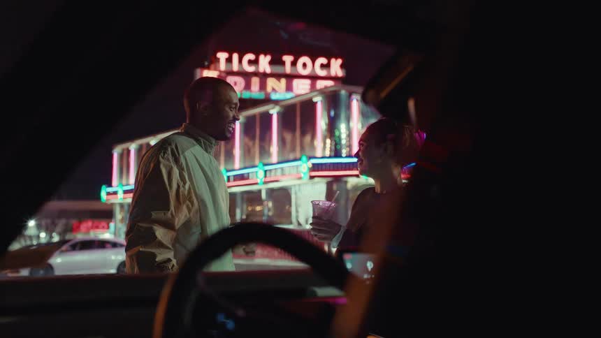 Couple smiles while standing in the parking lot of a diner at night and talking