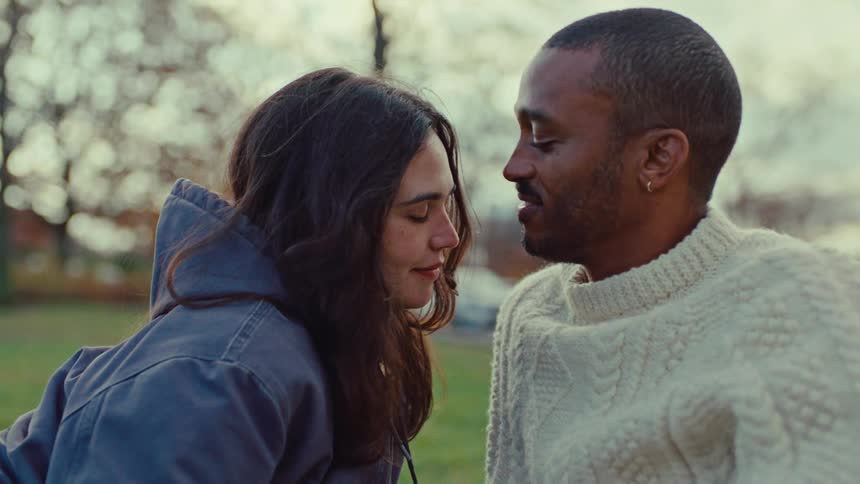 A man wearing a white sweater kisses a woman on the forehead while they sit together in a park during a close view