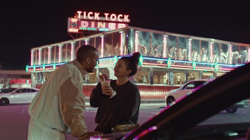 A man and woman stand by a car at night in front of a neon diner while the woman holds a milkshake and they laugh together in the parking lot
