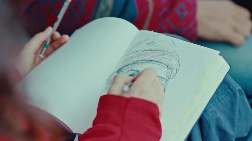A female sketch artist sitting in the passenger seat of a car drawing the face of a man sitting beside her at midday