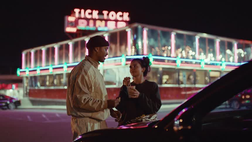Couple eats from a plate lying on the hood of a car while standing in the parking lot of a diner at night