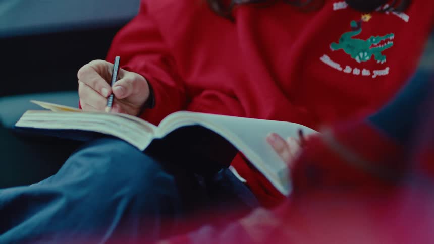 A child wearing a red sweatshirt sits and writes in a notebook with a black pen during a close view