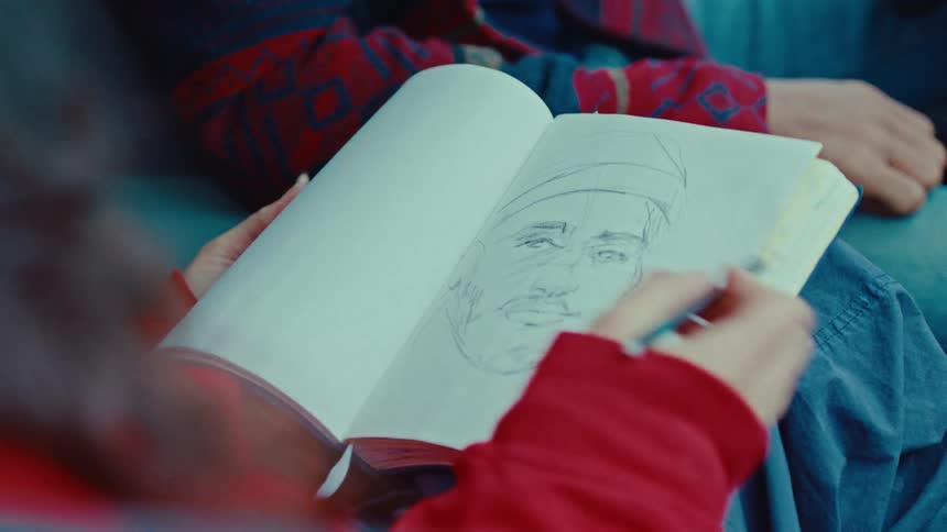 A girl wearing a red sweater draws a portrait of a person in a small notebook with a pencil