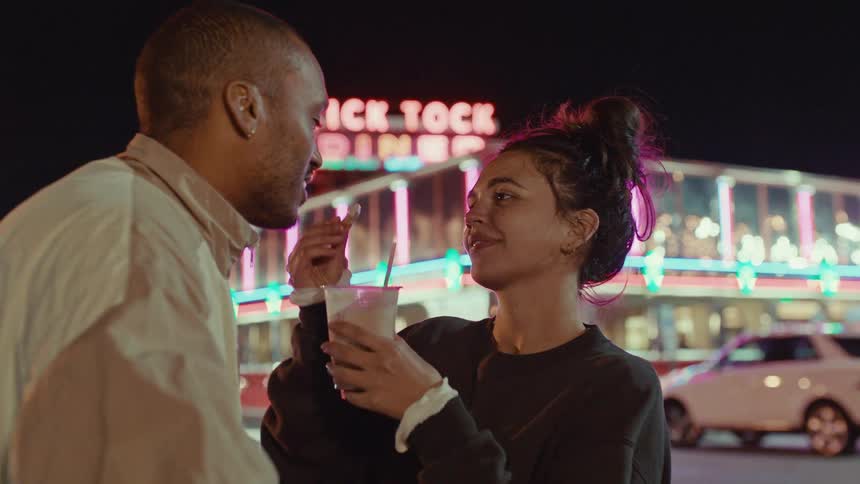 Woman offering dessert to a man in front of a diner with a neon sign at night