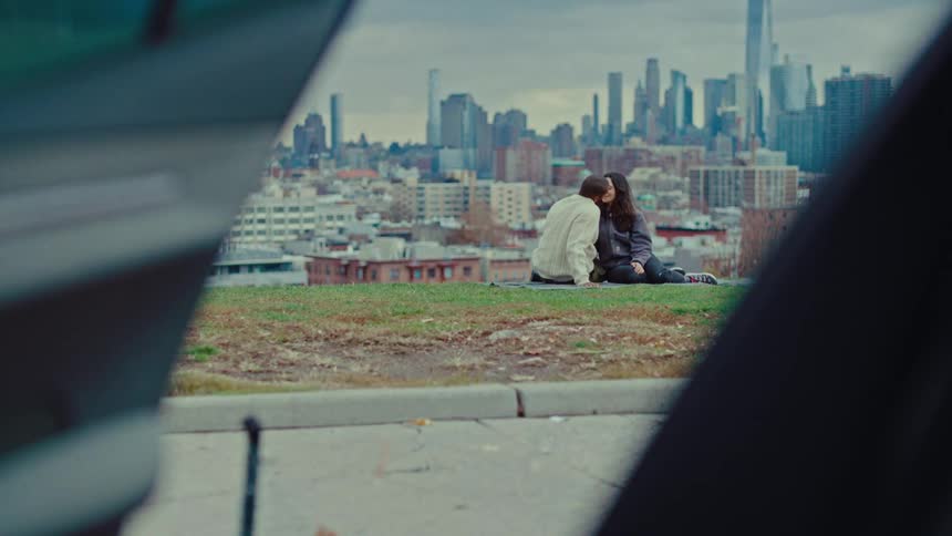 A man and woman sit together on a grassy hilltop overlooking a sprawling urban skyline leaning in for a gentle kiss as the hazy daylight illuminates the dense rows of skyscrapers and distant towers