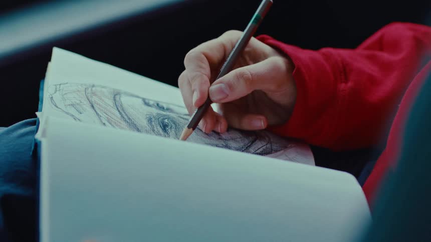 An artist sitting inside a car uses a pen to sketch a mans face at midday