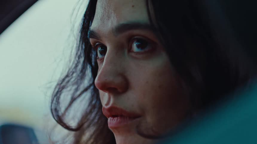 A woman with long dark hair looks out of a car window while sitting in the passenger seat