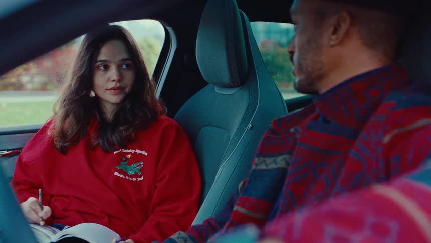 A young woman in a red sweatshirt sits in the passenger seat of a car drawing in a notebook while a man in a patterned jacket looks at her during a quiet moment parked outside