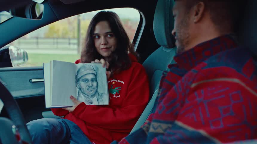 A young woman in a red hoodie sits in a car and happily shows a charcoal portrait she drew in a sketchbook to the man sitting in the driver seat next to her