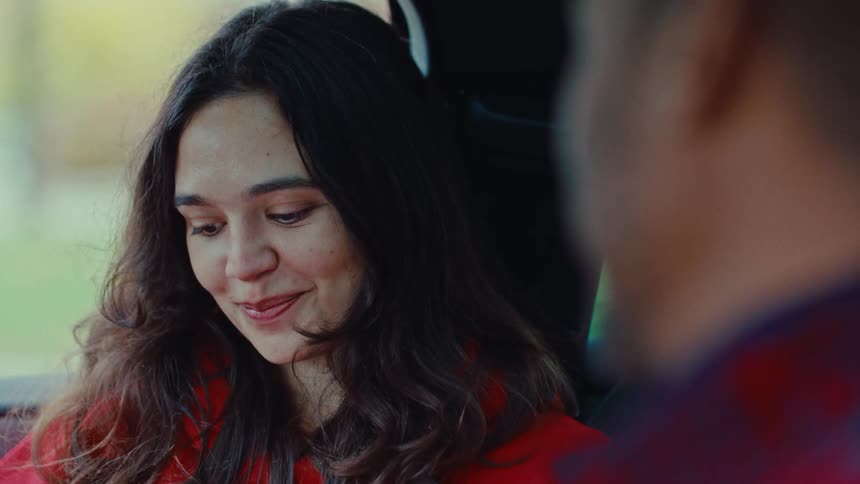 Woman with dark hair and a red coat smiles while looking at another person inside a car