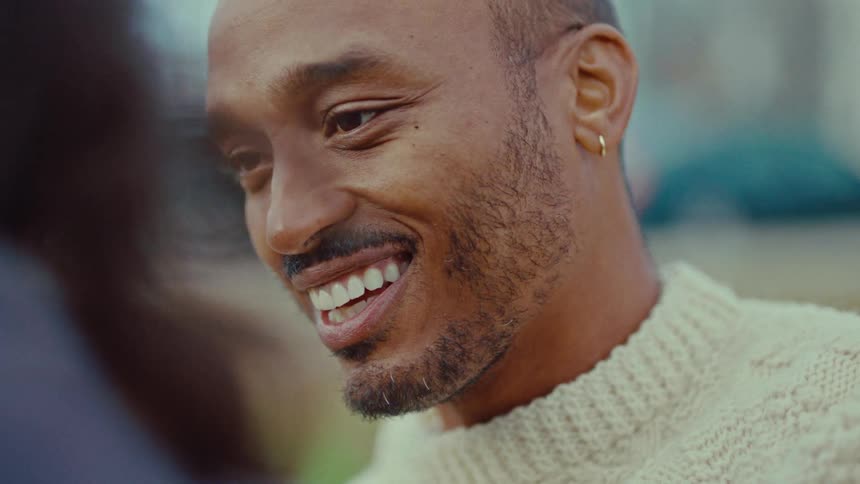 A man with a thin mustache and gold hoop earring smiles warmly while looking at someone offcamerahe wears a creamcolored cableknit sweater and eventually looks down with a bashful happy expression