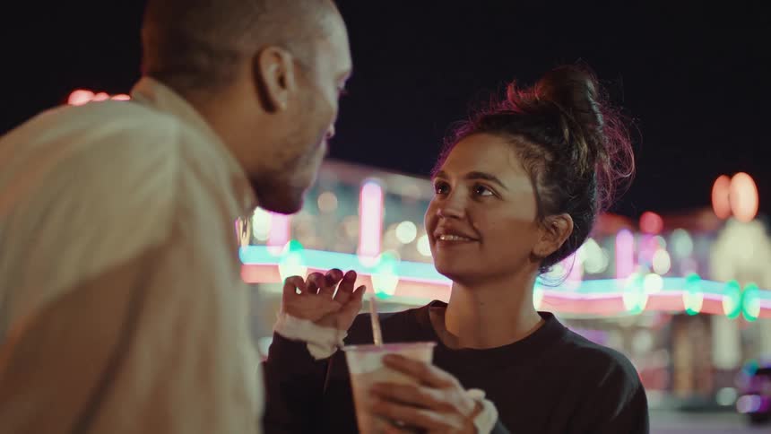 A young woman feeds a snack to a man while they stand together at a fair or carnival at nightshe smiles warmly at him after he takes a bite then takes a sip of her drink as they continue their conversation