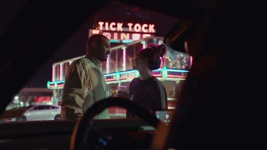 A man and woman stand outside a brightly lit tick tock diner at night talking and sharing a close momentthey lean in for a brief kiss before smiling at each other as they continue their conversation