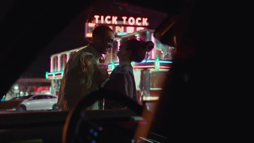 A man and a woman stand outside a brightly lit diner at night talking and laughing while the woman holds a drink and neon signs reflect on the parked cars nearby