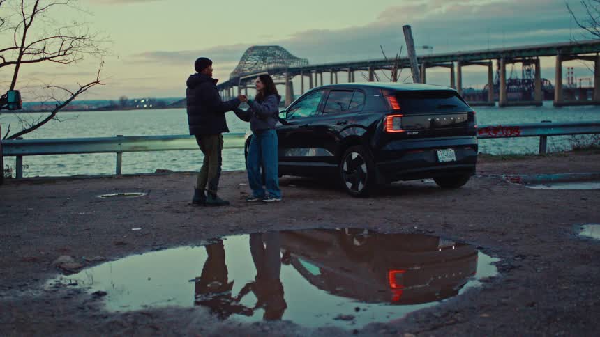 A couple stands together by a black suv parked near a body of water with a large bridge and city infrastructure visible in the background under a twilight skythe scene is reflected in a large puddle in the foreground capturing the pair a