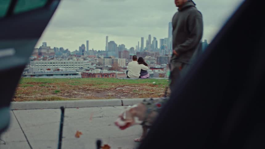 A man walks a dog on the sidewalk with a couple sitting and hugging in the background with a large skyline visible