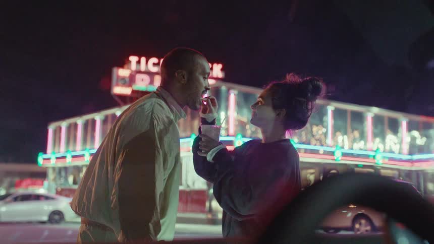 A man leans in closely toward a woman while they share a lighthearted smiling interaction outside a diner called tick tockthe woman holds a drink and playfully touches the mans face before they both turn their attention toward something in the distan
