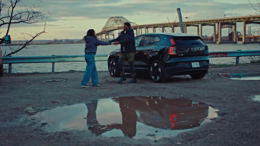 In a desolate parking lot by the water a young couple shares a slow intimate dance next to their black suvthe overcast sky and distant industrial bridge create a moody cinematic backdrop that is reflected in a large puddle at their feet