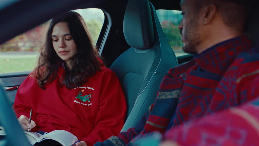 A woman in a red sweatshirt sits in the passenger seat of a car holding a pen and looking down at an open notebooknext to her a man in a patterned shirt sits in the drivers seat and looks toward her as they appear to be having a convers