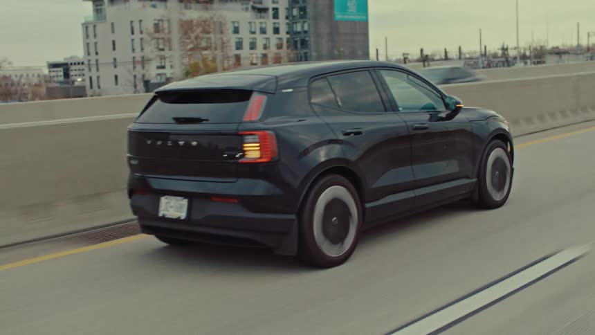 A black electric car drives along a concrete highway past modern buildings on a cloudy day
