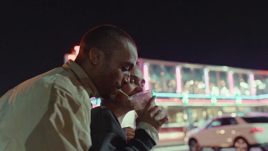 A man in a tan jacket leans in close to a smiling woman as she takes a sip of a milkshake through a straw at night with the bright neon lights of a vintage diner and a parked car blurred in the background