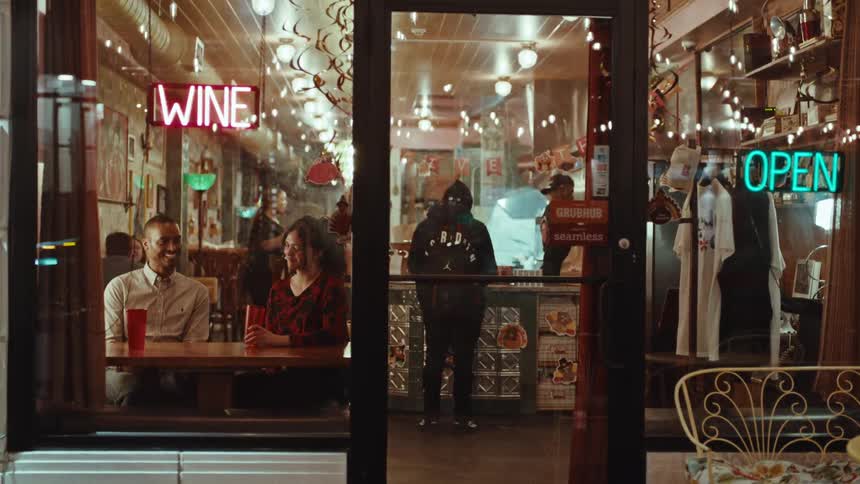 A man and a woman on a date sit at a table inside a brightly lit restaurant at night through a glass door