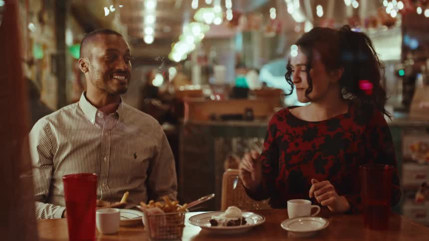 A man and a woman sit at a dimly lit restaurant table sharing fries and dessert they talk and laugh together while picking at the food in a relaxed cozy atmosphere