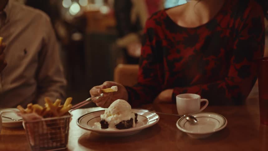 Young adult woman eating fries and ice cream sitting on a restaurant next to a man with other people walking on the background seen through a glass window during night time