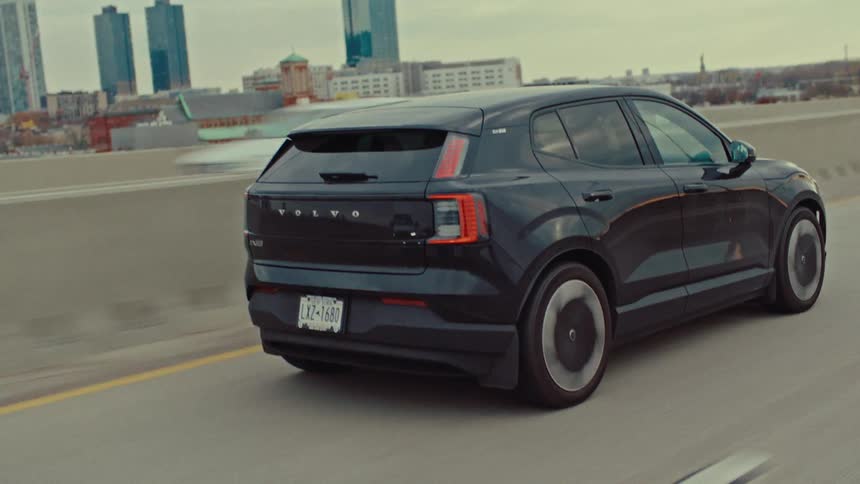 A black electric car drives along a concrete highway past modern buildings on a cloudy day