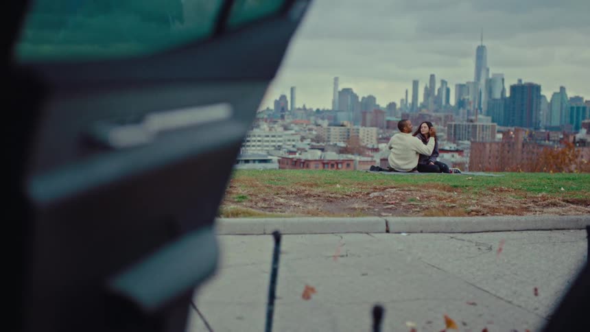 A man and a woman sit together on a grassy hill while looking at each other in front of a city skyline with tall buildings and a large tower under a cloudy sky