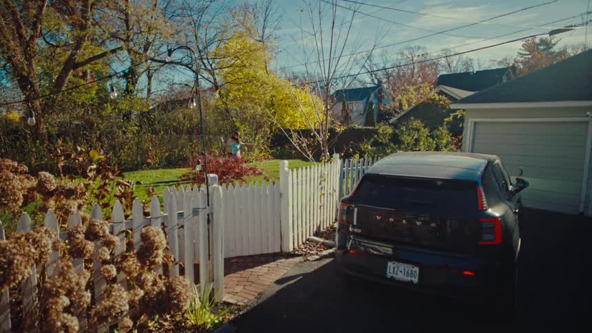Car parked in a neighborhood driveway next to a white picket fence as people in the yard do cartwheels