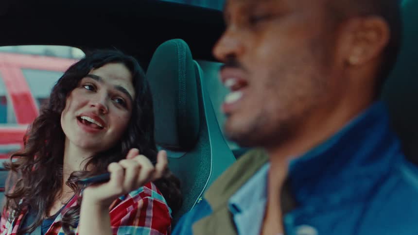 A woman in a red and white plaid shirt sings passionately while holding her phone like a microphone in the passenger seat of a carbeside her a man with a beard sits in the drivers seat smiling and joining in on the song as they drive together