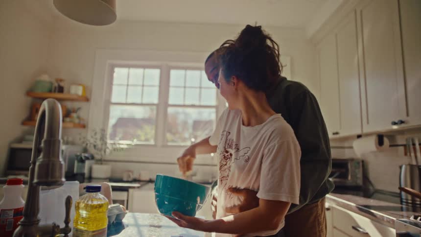 Man hugging a woman while she mixes batter in a bowl in the kitchen