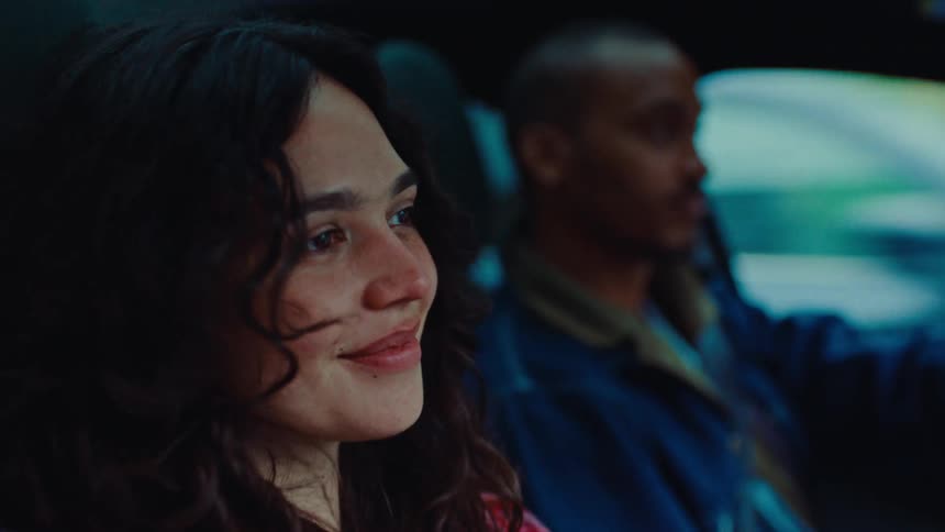 A young woman with curly hair smiles while sitting in the passenger seat of a car as a man drives through a city