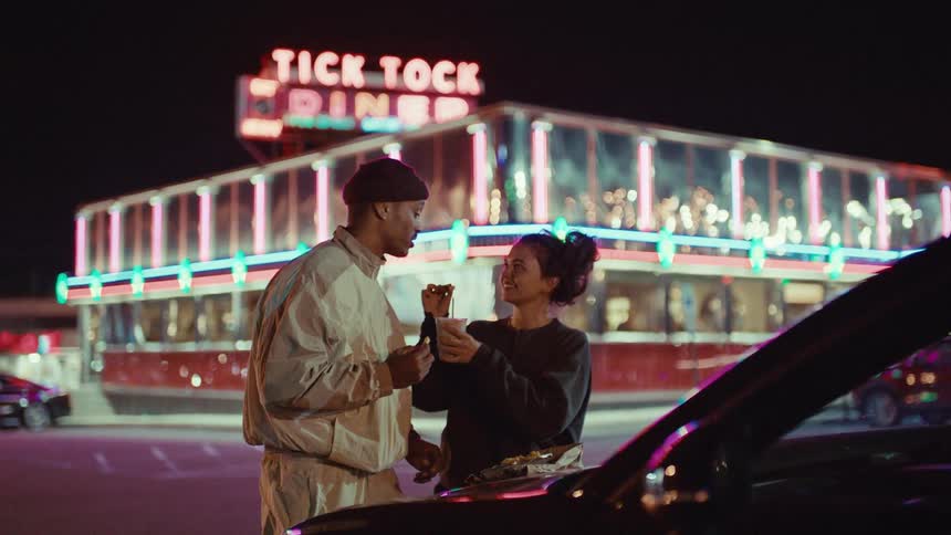 A man and woman eat fast food while standing outdoors next to their car at night