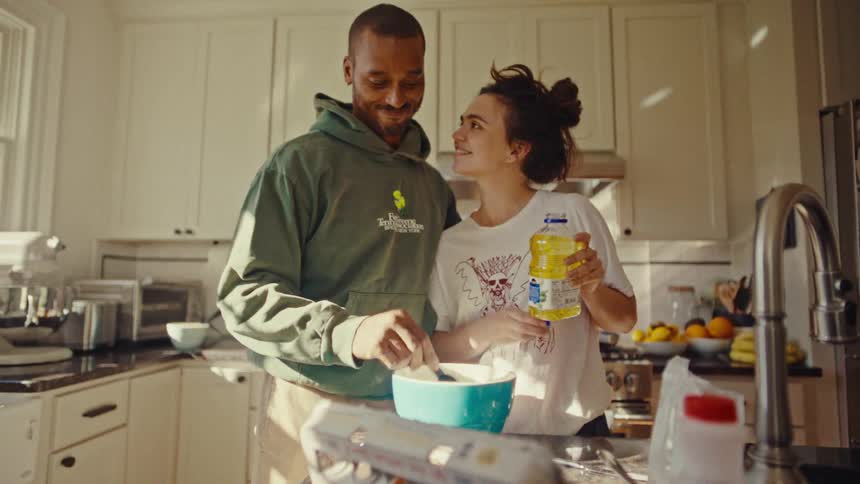 A man in a green hoodie and a woman in a white t shirt prepare food together in a bright kitchen as the woman pours oil into a measuring spoon held by the man